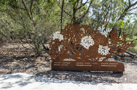 A large rusted cut out sculpture with native floral emblems form the local area. it is located in a bush setting along a creek.