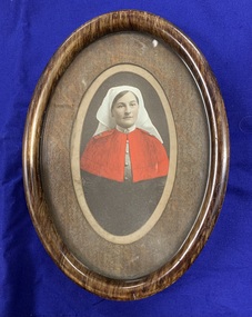 Brown wooden frame containing a hand-coloured photograph portrait of nurse Matron [Jean Matilda?] Stanbury, wearing a white wimple and red woolen cape.