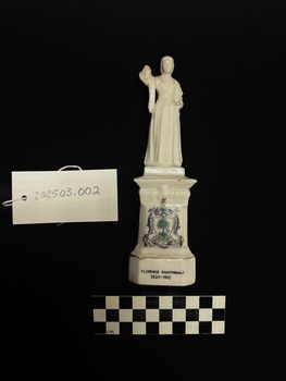 Photo of white ceramic Florence Nightingale statuette, with yellow, pink, blue, green and grey Glaswegian coat of arms and gold piping, laying flat upon a black background, scale card and object number also present. 