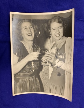 Black and white photograph of Margaret Rogers and Cath Burrows, Margaret laughing and Cath smiling, both holding a drink and wearing dresses adorned with medals, at a post World War Two function.
