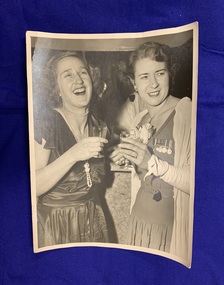 Black and white photograph of Margaret Rogers and Cath Burrows, Margaret laughing and Cath smiling, both holding a drink and wearing dresses adorned with medals, at a post World War Two function.