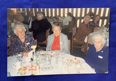 Colour photo of 3 older women sitting at a table at a function, with other tables of people in the background. 