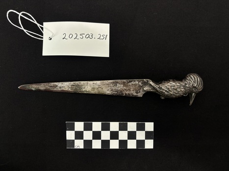 Photo of silver, chrome-plated letter knife, with kookaburra hilted handle, upon black fabric. On one of the blade faces the logo of the Adelaide Steamship Company, a white flag, with a blue cross and an eight-pointed red star in the centre. On the blade face accompanying the flag the ship name, M.V. Manunda, is also pressed into the blade. On the opposing face of the blade, a makers mark and silver quality mark is present. Scale card and object number also present. 