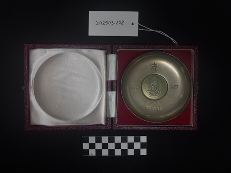 Photo of Queen Elizabeth II Silver jubilee dish laid upon the dish's matching red felt box. The cool-to-touch silver dish has engravings upon its face including the royal mark of Queen Elizabeth the Second, 'EIIR' under surmounted by St Edward's crown, '1952' on the left of the dish, 'Royal Australian Army Nursing Cross' is embossed within an oval central press, surmounted by St Edwards crown, in the centre of the oval is a Nightingale oil lamp, across the bottom of the central oval is a scroll embossed with the motto 'Pro Humanitate' Latin for 'For Humanity'. Underneath the central embossment and aligned with the royal Elizabeth sigil is an engraving of the dates '1902 - 1977' [seemingly this date was engraved after the making of the dish, it is unknown how long after the presenting of the dish the engraving was added.] Underneath those dates are engravings/marks of silver, nickel and makers quality marks. Upon flipping the dish over, on the slight foot of the dish is an embossing of 'Nickel Silver' and an engraving of the silver quality number '49'. The red felt box lid is also pictured, white satin with soft cushioning to protect the dish when closed. This photograph is taken upon a black background, with a scale card and object number included.