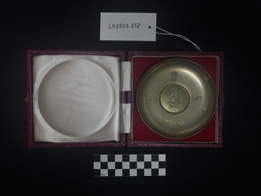 Photo of Queen Elizabeth II Silver jubilee dish laid upon the dish's matching red felt box. The cool-to-touch silver dish has engravings upon its face including the royal mark of Queen Elizabeth the Second, 'EIIR' under surmounted by St Edward's crown, '1952' on the left of the dish, 'Royal Australian Army Nursing Cross' is embossed within an oval central press, surmounted by St Edwards crown, in the centre of the oval is a Nightingale oil lamp, across the bottom of the central oval is a scroll embossed with the motto 'Pro Humanitate' Latin for 'For Humanity'. Underneath the central embossment and aligned with the royal Elizabeth sigil is an engraving of the dates '1902 - 1977' [seemingly this date was engraved after the making of the dish, it is unknown how long after the presenting of the dish the engraving was added.] Underneath those dates are engravings/marks of silver, nickel and makers quality marks. Upon flipping the dish over, on the slight foot of the dish is an embossing of 'Nickel Silver' and an engraving of the silver quality number '49'. The red felt box lid is also pictured, white satin with soft cushioning to protect the dish when closed. This photograph is taken upon a black background, with a scale card and object number included.