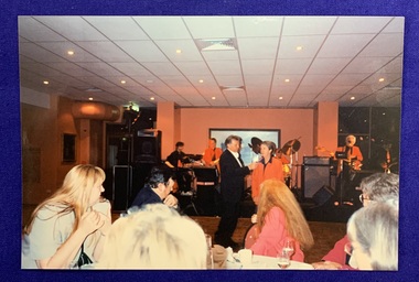 A group of people in the foreground look towards the dancefloor where Normie Rowe is holding a microphone towards Jan McCarthy who is speaking. A 4 or 5 piece band can be seen in the background. 
