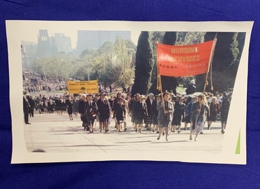 A pixelated colour photograph of Returned Nurses marching in the forecourt of the Shrine of Remembrance with the Melbourne central business district in the background. Some are holding banners that say Nursing Services R.A.N.N.S. R.A.A.F.N.S. and the Australian Women's Army Service, Australia New Guinea Victoria.