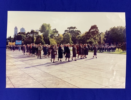 A large colour photograph of Returned Nurses marching in the forecourt of the Shrine of Remembrance (Melbourne, Victoria) of ANZAC Day. Women in uniform are marching and saluting, there is a large marching band playing in the background.