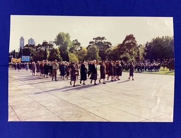 A large colour photograph of Returned Nurses marching in the forecourt of the Shrine of Remembrance (Melbourne, Victoria) of ANZAC Day. Women in uniform are marching and saluting, there is a large marching band playing in the background.