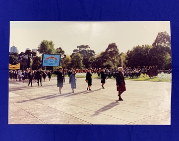 A large colour photograph of Sister Elizabeth Burchill and four physiotherapists marching in the forecourt of the Shrine of Remembrance on ANZAC Day in 1991. More Returned Service League (RSL) Members march in the background carrying banners, and there is a large marching band playing.
