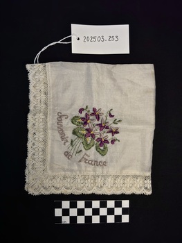 Photograph of silky, cotton handkerchief with lace trim and purple, green, yellow and mauve embroidery depicting a bouquet of flowers [either iris', the national flower of France or lilacs symbolising innocence, death and mourning.]. The photograph is taken upon a black background with scale card and object number present. 