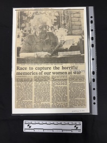 Newspaper clipping with four columns of text, headed by a large black and white photograph of an elderly woman sitting at a table looking at documents
