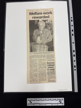 Newspaper clipping with a headline and large black and white photograph of a elderly woman holding an award in her lap.  The article has three columns of text.