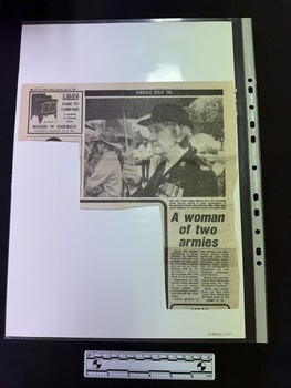 Newspaper clipping with advertisement in top left hand corner, first part of article has two columns with a large black and white photograph of an elderly woman in uniform holding an umbrella.  Article continues on a page not attached