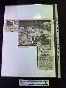 Newspaper clipping with advertisement in top left hand corner, first part of article has two columns with a large black and white photograph of an elderly woman in uniform holding an umbrella.  Article continues on a page not attached