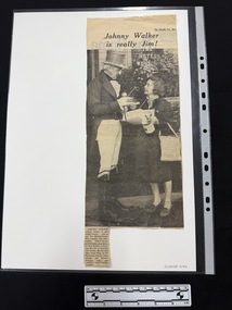 Newspaper clipping of a large black and white photograph of an older man in a top hat talking to a shorter woman holding a collection tin.  The photograph has a caption and heading.