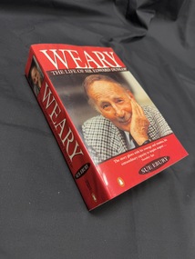Paperback book with a red cover that has a colour photo of an elderly man on the cover and spine, and white writing on the cover and spine