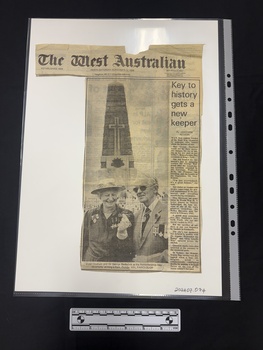 Clipping has masthead and article with long photograph of older man and woman standing infront of an obelisk shaped memorial and one column of text.