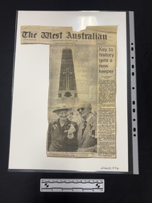 Clipping has masthead and article with long photograph of older man and woman standing infront of an obelisk shaped memorial and one column of text.