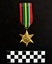 Photo of the front of a The 1939-1945 Star medal. Made from yellow copper zinc alloy, the medal itself is cool-to-touch, with somewhat sharp 6 points. The obverse has a central and textured design of the Royal Cypher 'GRJ VI', surmounted by a crown. A circlet, the top of which is covered by the crown, surrounds the cypher and is inscribed 'THE 1939 - 1945 STAR'. The ribbon is tactile, feeling ribbed under finger. This photograph is taken upon a black background, with a scale card and object number included. [It appears the ribbons from this medal and 202508.260 have been added to the wrong medal.]