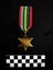 Photo of the front of The 1939-1945 Star medal. Made from yellow copper zinc alloy, the medal itself is cool-to-touch, with somewhat sharp 6 points. The reverse is engraved with 'VFX53656 J.G. GIBSON'. The ribbon is tactile, feeling ribbed under finger. This photograph is taken upon a black background, with a scale card and object number included. [It appears the ribbons from this medal and 202508.260 have been added to the wrong medal.]