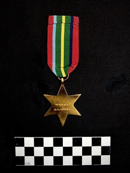 Photo of the front of The 1939-1945 Star medal. Made from yellow copper zinc alloy, the medal itself is cool-to-touch, with somewhat sharp 6 points. The reverse is engraved with 'VFX53656 J.G. GIBSON'. The ribbon is tactile, feeling ribbed under finger. This photograph is taken upon a black background, with a scale card and object number included. [It appears the ribbons from this medal and 202508.260 have been added to the wrong medal.]