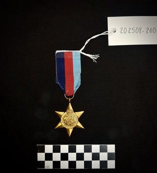 Medal from yellow copper zinc alloy with somewhat sharp six points. The obverse has a central and textured design of the Royal Cypher 'GRJ VI', surmounted by a crown. A circlet, the top of which is covered by the crown, surrounds the cypher and is inscribed 'THE PACIFIC STAR'. The ribbon is tactile with red, navy, and light blue stripes. [I believe that the ribbon on 202508.259 and 202508.260 have been swapped at some point.]