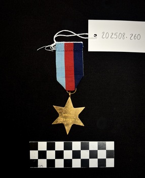 Photo of the reverse of a Pacific Star medal. Made from yellow copper zinc alloy, with somewhat sharp 6 points. The reverse 'VFX53656 J.G. GIBSON' engraved on it. The ribbon is tactile with red, navy, and light blue stripes.  [I believe that the ribbon on 202508.259 and 202508.260 have been swapped at some point.]
