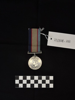 Photo of the reverse view of the Australia Service Medal. Made from nickel-silver, the reverse has a central and textured design of the crowned effigy of King George VI, facing left and signed 'PM', the initials of designer Percy Metcalfe, below the truncated neck of the effigy. Around the perimeter is the legend 'GEORGIVS VI D:G:BR:OMN:REX ET INDIAE IMP:' The ribbon is tactile, feeling ribbed under finger. This photograph is taken upon a black background, with a scale card and object number included.