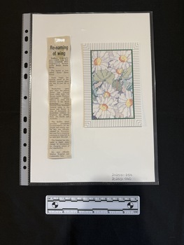 A card with a picture of colorful flowers, framed by stripes, sitting to the right of a newspaper clipping with a headline and single column of text