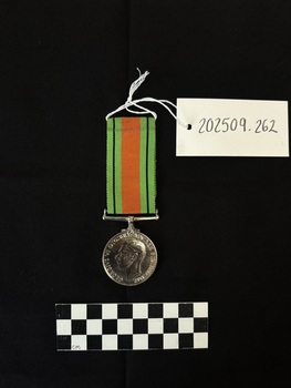 Photo of the obverse of a Defence Medal. Made from cupro-nickel, the medal itself is cool-to-the-touch. The obverse has a central and textured design of the uncrowned effigy of King George VI, facing left and signed 'HP', the initials of designer T.H. Paget, below the truncated neck of the effigy. Around the perimeter is the legend 'GEORGIVS VI D:G:BR:OMN:REX ET INDIAE IMP:' The rim of the medal has two engravings: 'VFX53656/J.G. GIBSON'. The ribbon is tactile, feeling ribbed under finger. This photograph is taken upon a black background, with a scale card and object number included.