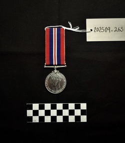 Front view of the War Medal 1939-1945. The silver coloured medal has a central and textured design of the crowned effigy of King George VI, facing left, around the perimeter is the legend 'GEORGIVS VI D:G:BR:OMN:REX ET INDIAE IMP:' The ribbon is coloured in vertical strips: red, blue, white, red, white, blue, red, it is tactile, feeling ribbed under finger. This photograph is taken upon a black background, with a scale card and object number included.