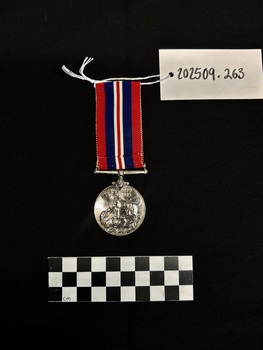 Back view of the War Medal 1939-1945. The silver coloured medal has a lion standing on a double-headed dragon. The ribbon is coloured in vertical strips: red, blue, white, red, white, blue, red, it is tactile, feeling ribbed under finger. This photograph is taken upon a black background, with a scale card and object number included.