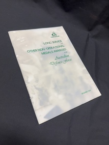 Staple bound booklet with opaque cover containing green metallic writing over card cover collage of photographs of military persons and equipment