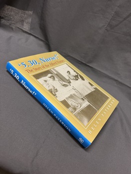 Hardback book with olive green, blue, red and white cover and writing.  Cover has sepia image of two nurses working in a hospital ward, one talking to a patient in a bed.