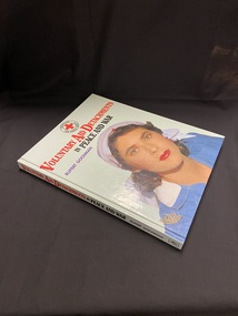 Hardcover book with a light green cover. The Title is printed in red and black. There is a female nurse on the front cover in a blue uniform and white red cross head scarf. The Australian Red Cross symbol is at the top of the front cover.