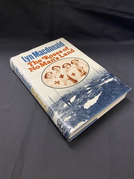 Hardcover book with dust jacket. Title is brown print. Author's name in dark green print above the title (although it appears blue in image). There is a sepia photo of four nurses wearing Red Cross uniforms that overlays a black and white background image of a soldier walking through muddy land with  burnt tree trunks behind him.