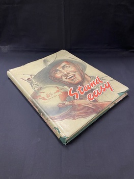 Green hardcover book with title in lighter green writing on the front cover and spine and an image in the same light green of a soldier holding a rifle with his arms held up in victory. There is a worn and slightly torn pale olive dustjacket on the book, it has 'Stand easy' written on the lower left side of the front cover in red print. There is a large illustration of a soldier's head and upper body, he is smiling and holding a world globe (with Australia clearly visible) in the palm of his hand. On top of the globe stands another soldier holding a rifle with his arms held high in triumph. The edges and spine of the dust jacket are very torn and the writing on the spine is quite faded.