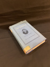 Navy blue hardcover book with lighter blue dust jacket. The front cover has a black ornate border around the title (printed in white) and the subtitles and author's name printed in black. In the centre of the cover is a black pen sketch of a man in an oval shape. At the top of the spine is a cream coloured square with the main title printed in black. The subtitle and author details are on the blue part of the spine, also printed in black. At the bottom of the spine is an old catalogue number on a sticker that has been covered in tape, which has yellowed with age.