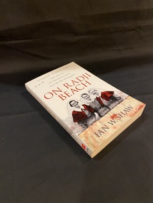 White paperback book. At the top of the cover are three small black planes flying to the left of the sub-heading. Below this is the title printed in large red letters and underneath is a picture of three smiling Australian nurses standing close together, the two nurses at either end have red capes and the rest of the photo is in black and white.
Directly below the image is a line of black barbed wire and at the bottom of the page is a feint image of soldiers drawing fire towards a ship and the red Japanese sun symbol overlayed across the top of the image.