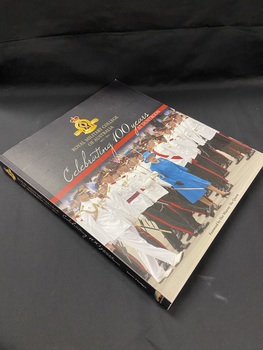 Large paperback book with a black cover and a photo of the Queen in a blue dress walking in front of soldiers standing in formation in white dress uniforms. The top third of the cover has a black panel with the title printed below the emblem for the Royal Military College. At the bottom of the panel is a red stripe with 'AT DUNTROON' printed in white on the right hand side.