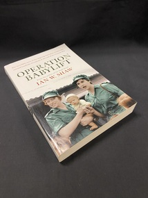 White book with a picture of two women in green uniforms, one of whom is holding a baby, in front of a building with a corrugated roof. The title 'OPERATION BABYLIFT' is printed above the image in green, followed by the author's name in red text. At the top of the page it reads "The incredible story of the inspiring Australian women who rescued hundreds of orphans at the end of the Vietnam War".