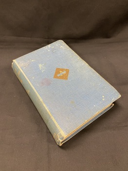 Faded blue cloth bound, hardcover book. The title is printed down the spine in gold letters. There is a small, gold, diamond shape in the middle of the cover with an empty rectangle inside it and the letter A embossed in it.