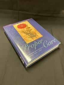 Hardback book with a dark blue dustjacket. Along the spine the title is printed in light aqua script, followed by the printed text of the sub-title and author's name, there is a mark on the bottom of the spine where a sticker has previously been removed.
The front shows an image of a red flower with some greenery set in a light brown rectangle, below which the title is again printed in light aqua script.