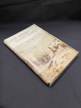 Black hardcover book with a dustjacket. Sepia coloured image of men riding a large group of camels through the desert wraps around the front and spine of the book. The title is printed in brown text across the top of the image and down the spine, over the image.