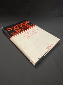 Grey dustjacket with a small tear on the bottom on a black hardcover book. There is a black panel around the top that wraps around the spine with the totle printed in red across it. Below this is a white and red stripe. The rest of the cover is grey with the subtitle and author's name printed in red print.
There is a white sticker attached to the bottom of the spine with B62 written on it.