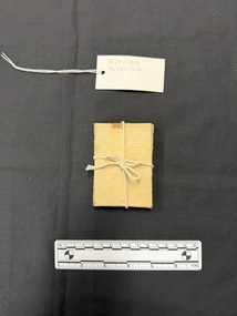 Small light brown cardboard box tied with natural coloured cotton string. This photograph is taken upon a black background, with a scale card and object number included.