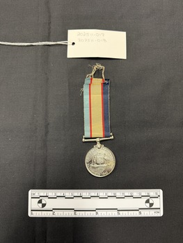 Photo of the obverse of a Australia Service Medal. Made from nickel-silver, the obverse has a central and textured design of the Australian coat of arms. The face has 'THE AUSTRALIA SERVICE MEDAL/1939-1945' inscribed. The rim of the medal is engraved with 'QF140158 M. L. JACKSON'. The ribbon is tactile, feeling ribbed under finger. This photograph is taken upon a black background, with a scale card and object number included.