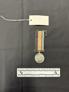 Photo of the reverse view of the Australia Service Medal. Made from nickel-silver, the reverse has a central and textured design of the crowned effigy of King George VI, facing left and signed 'PM', the initials of designer Percy Metcalfe, below the truncated neck of the effigy. Around the perimeter is the legend 'GEORGIVS VI D:G:BR:OMN:REX ET INDIAE IMP:' The ribbon is tactile, feeling ribbed under finger. This photograph is taken upon a black background, with a scale card and object number included.