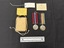 Small cardboard box with natural coloured string alongside two silver medals with coloured ribbed ribbons.  Also shown are two pieces of paper, one yellow with black type on it, the other piece a buff coloured heavier stock (card) with dark blue type and a handwritten note in the top left corner in blue ink.  This photograph is taken upon a black background, with a scale card and object number included.